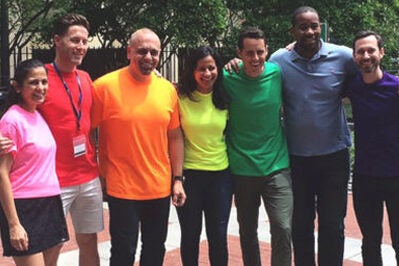 A group of people standing together outdoors, each wearing a different color of the rainbow, suggesting celebration and diversity.