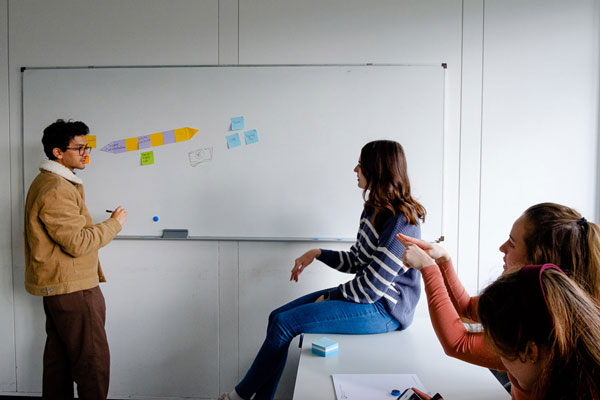 Ein Mann steht an einem Whiteboard auf dem Notizzettel kleben und diskutiert mit drei Frauen, die interessiert auf das Whiteboard schauen