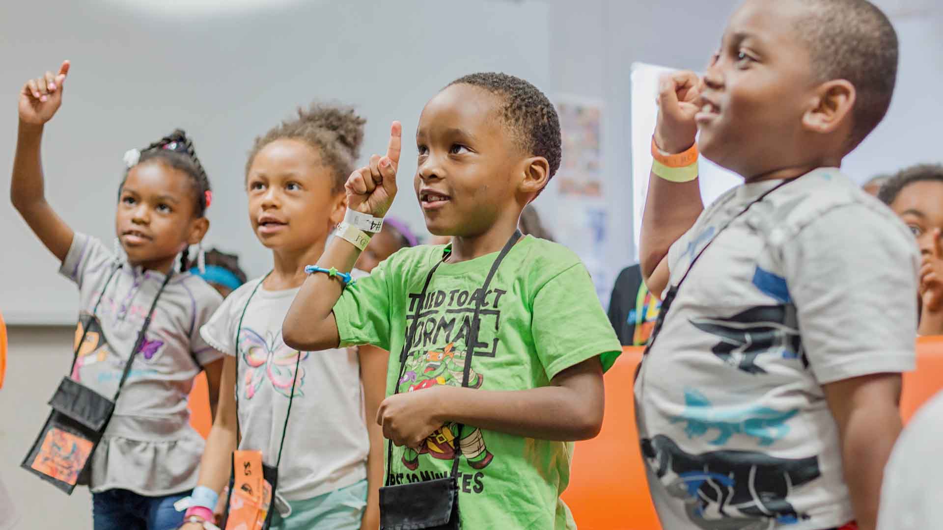 Four little kids at a summer camp. They're wearing brightly colored shirts and bracelets and several are raising a hand and index finger. 