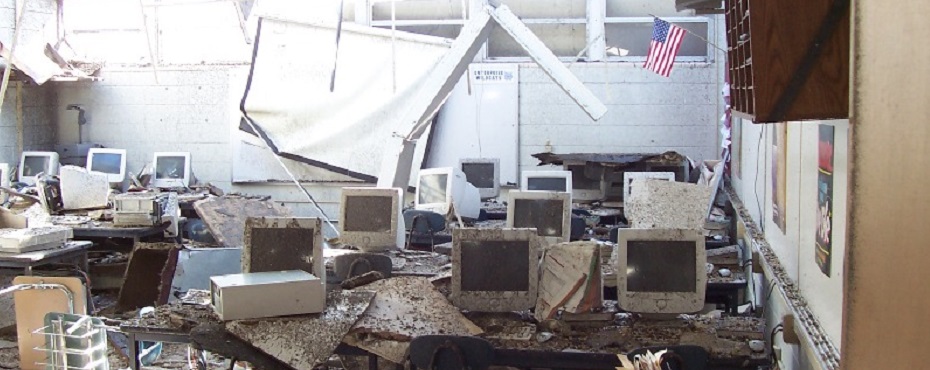 Computer Lab with tables and monitors covered in dust and debris. American Flag still hanging on wall.