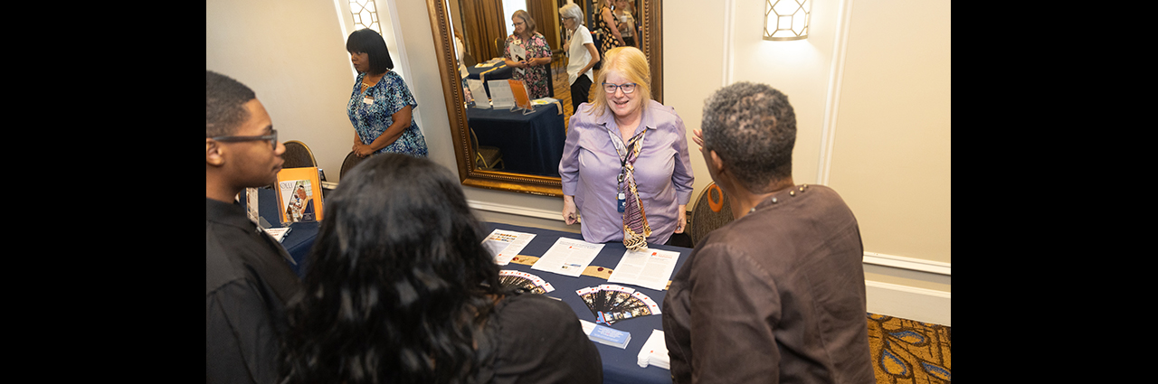 Two female staff members behind a table presenting flyers to several event attendees.