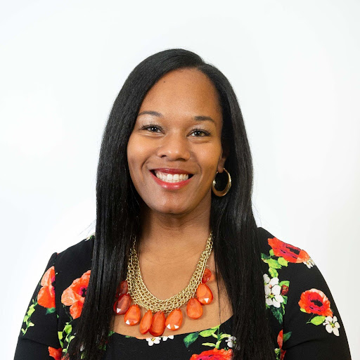 A medium-brown-skinned Black woman with straight black hair just past her shoulders is wearing a black dress with magenta flowers and green leaves, gold hoop earrings, and gold necklace with orange teardrop gems. She is smiling at the camera with a white background.