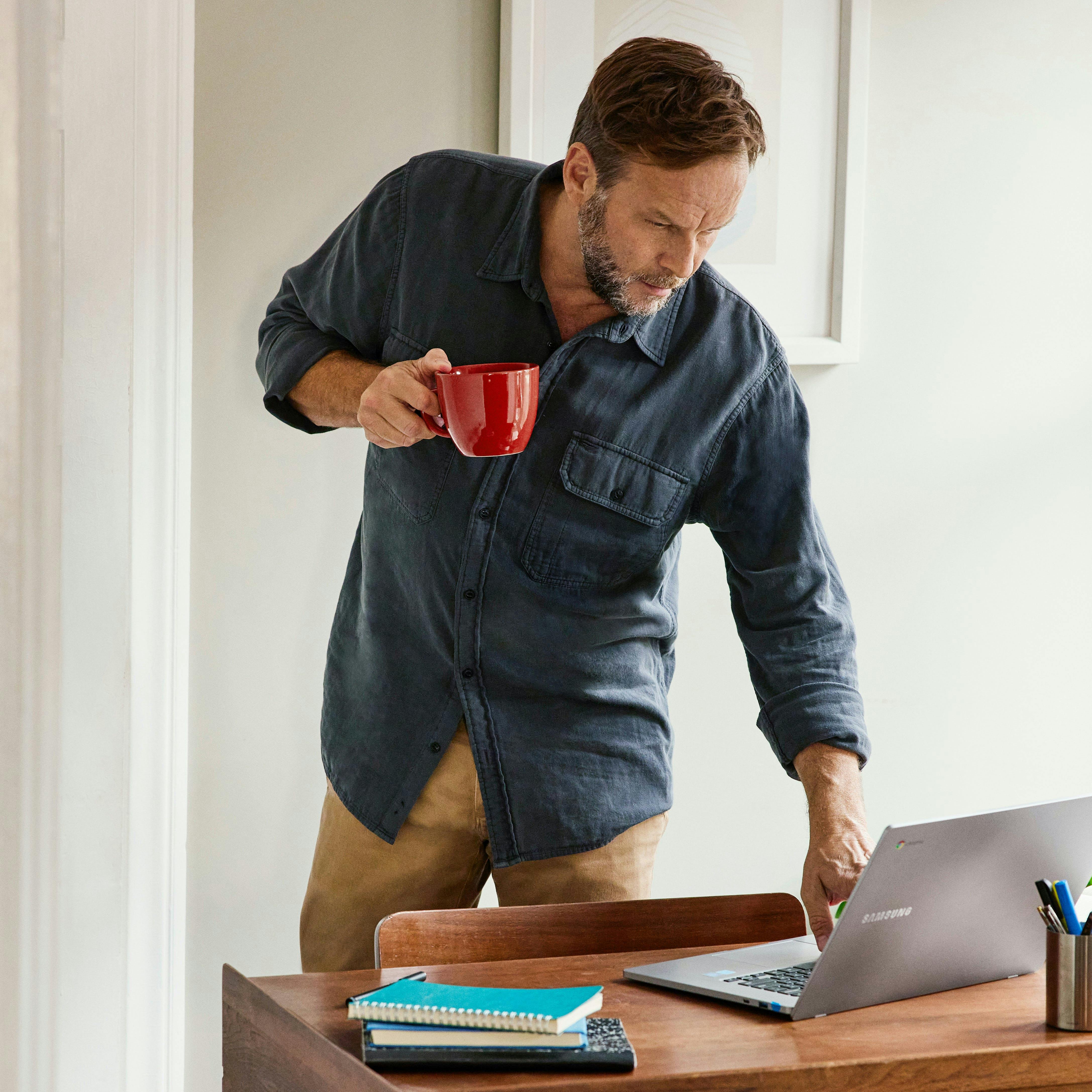 Man holding a red mug standing at a low desk touching pointing to the screen of a Chromebook