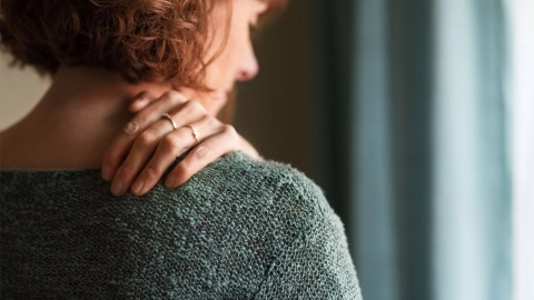 Eine Frau fasst sich mit der linken Hand an die rechte Schulter nahe des Halses. 