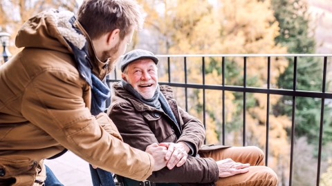 Ein im Rollstuhl sitzender älterer Mann und ein jüngere Mann bei einem Spaziergang. Der jüngere Mann stützt sich auf die Lehne des Rollstuhls, lehnt sich in Richtung des älteren Mannes und legt die Hand auf dessen Arm.