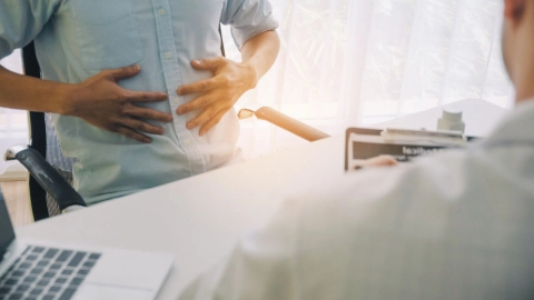 Ein Mann sitzt in einem Behandlungszimmer vor einem Arzt und fasst sich mit beiden Händen an den Bauch. Der Arzt hält einen Auswertungsbogen in der Hand.