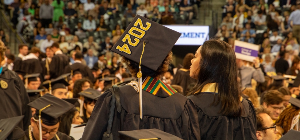 Students in their grad caps and gowns at the Spring 2024 commencement ceremony, with one wearing a gad cap with "2024" painted on it in gold.