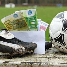 Ein Briefumschlag mit Geldscheinen steckt zwischen FuÃballschuhen, die auf der Auswechselbank eines Sportplatzes stehen.