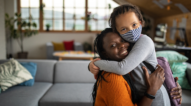 In a living room space, a Black mother warmly smiles and hugs her child