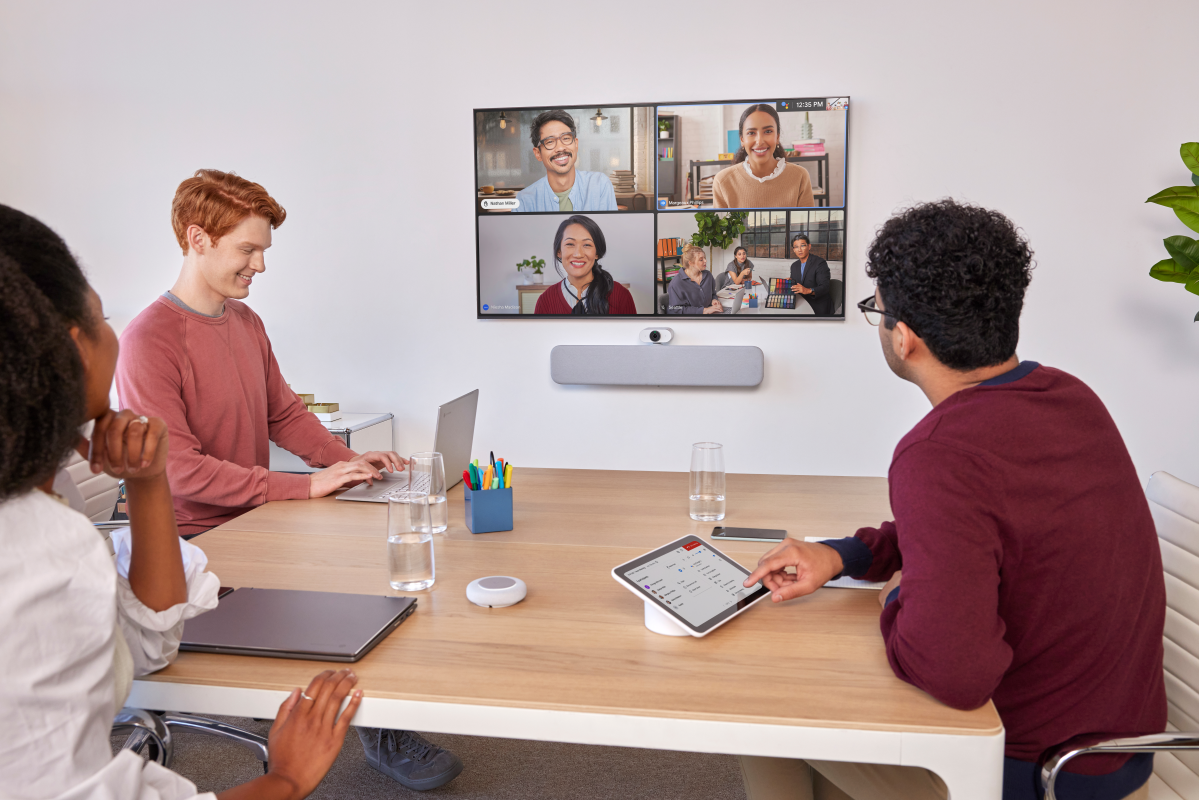 A meeting room with three people, and four shown on-screen in video conference