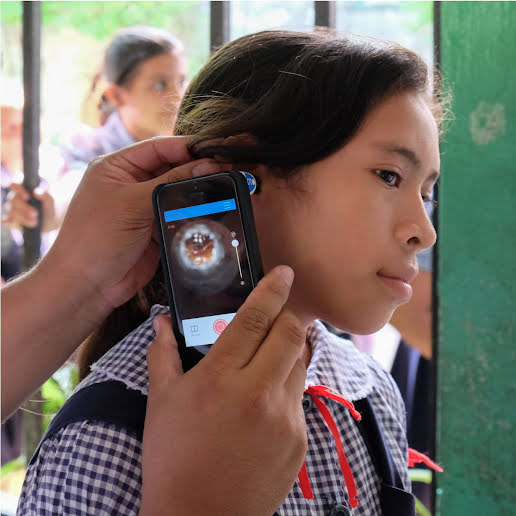 A healthcare worker uses a smartphone to examine a young girl's ear with an otoscope.