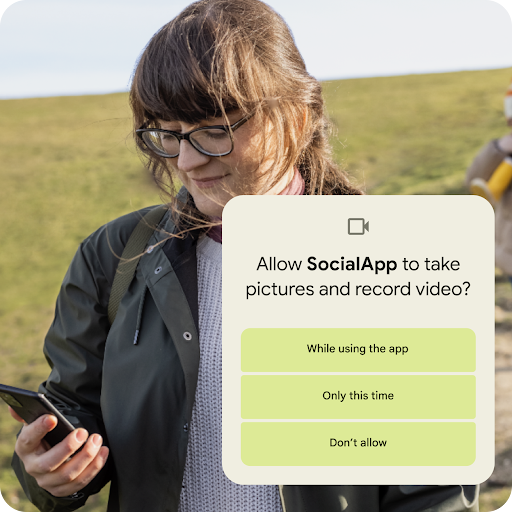 A person standing on a grassy hillslope looking at their Android phone. A graphic overlay seeking permission to allow an app to take pictures and record videos is placed on top. Permission options include allowing access while using the app, only this time or don't allow.