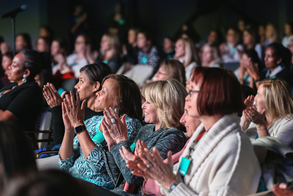 A group of women sit in a theatre and clap while looking at the stage