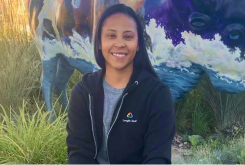 Maria, an Indigenous Googler, sits smiling in front of a buffalo sculpture, wearing a Google Cloud sweatshirt