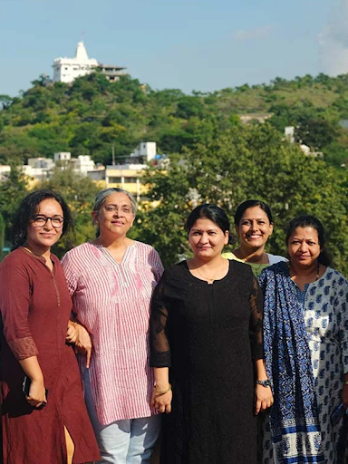 Five women wearing traditional Indian dresses stand smiling in front of a lush green landscape