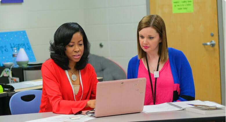 Two people sit at a desk and engage with a laptop.