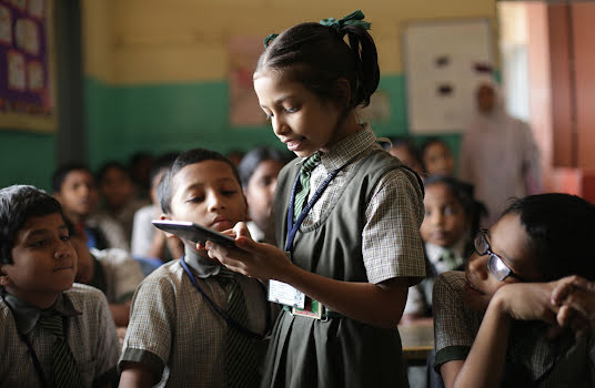 A young girl holding a tablet while classmates crowd around.