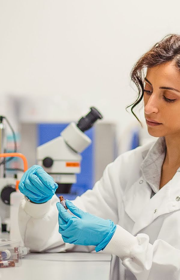 Woman in science lab wearing lab coat and medical gloves inspecting vial.