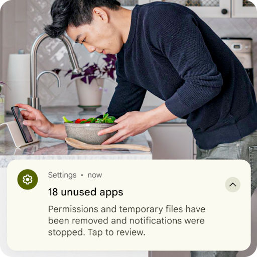 A person is preparing food by the kitchen sink while looking at their Android phone. A graphic overlay of a settings notification is placed on top of the image. It states that temporary files from unused apps have been removed and permissions have been reset.