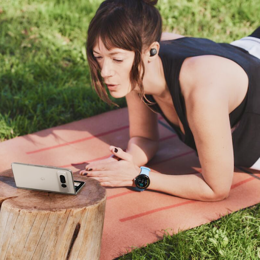 En person trener på en yogamatte, bruker en Wear OS-smartklokke og øreplugger og ser på en foldbar Android-telefon.