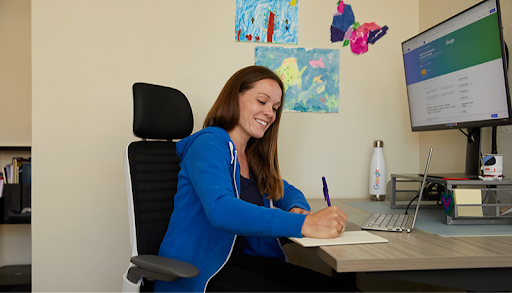 A woman in a home office space writes on a notepad. Childrens drawings can be seen on the wall behind her