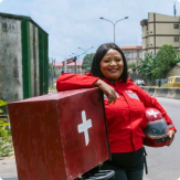 Une femme portant une veste rouge se tient debout sur un trottoir, appuyée sur une moto rouge, et tenant un casque.