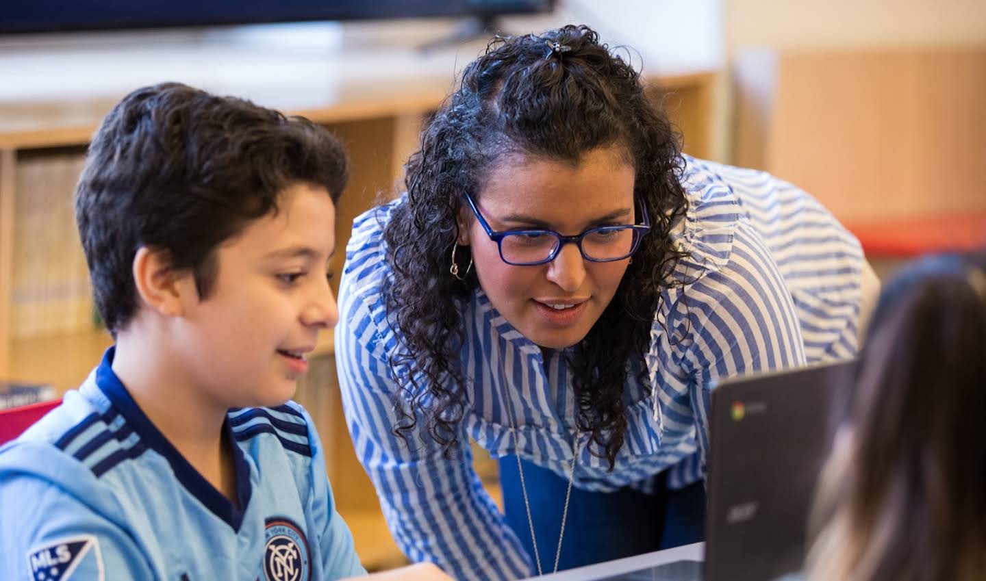 Primer plano de una educadora con gafas que se acerca para revisar algo en la pantalla de un alumno.