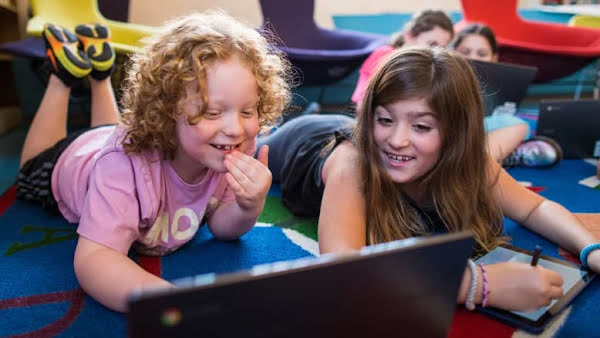 Two smiling children in a classroom learning on a laptop together.