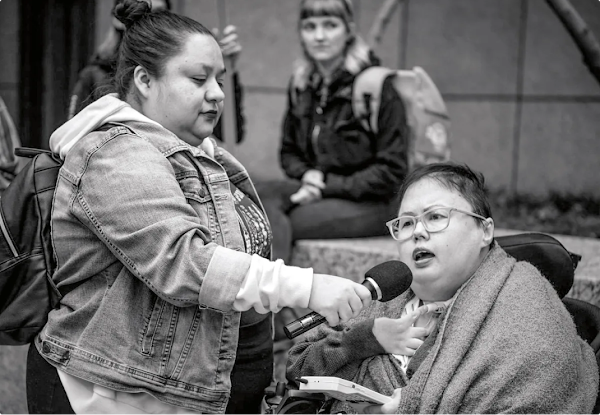 Une femme en fauteuil roulant parle dans un micro tenu par une femme debout à ses côtés.