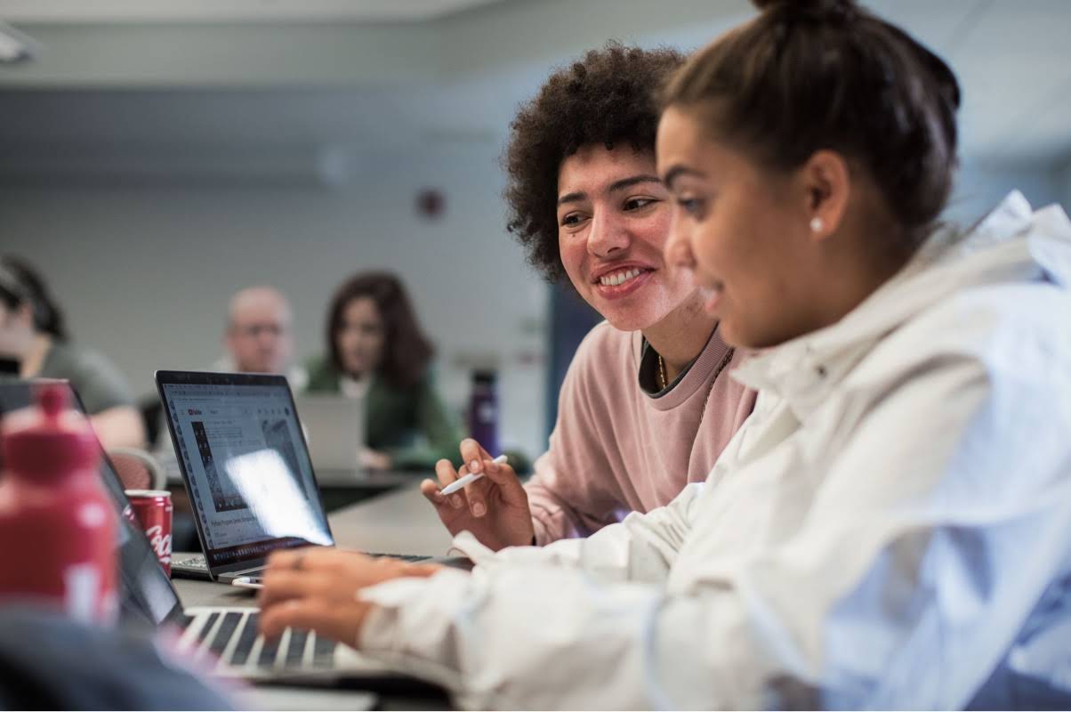 Una adolescente escribe en una computadora mientras recibe consejos de una instructora que tiene a su lado