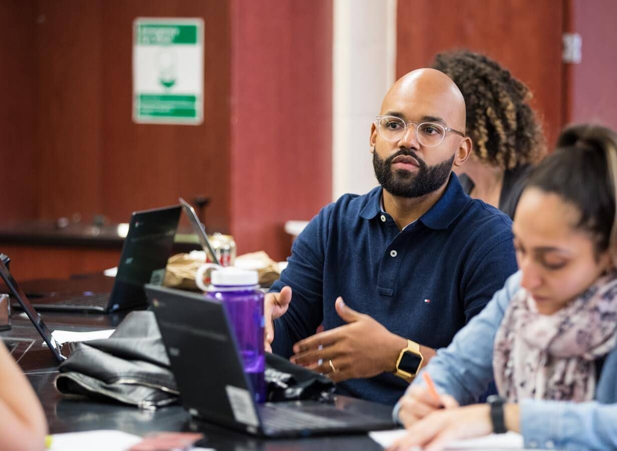 Homme assis dans une salle/salle de classe, le regard dirigé vers l'avant, et deux autres personnes à proximité travaillant sur des Chromebooks.