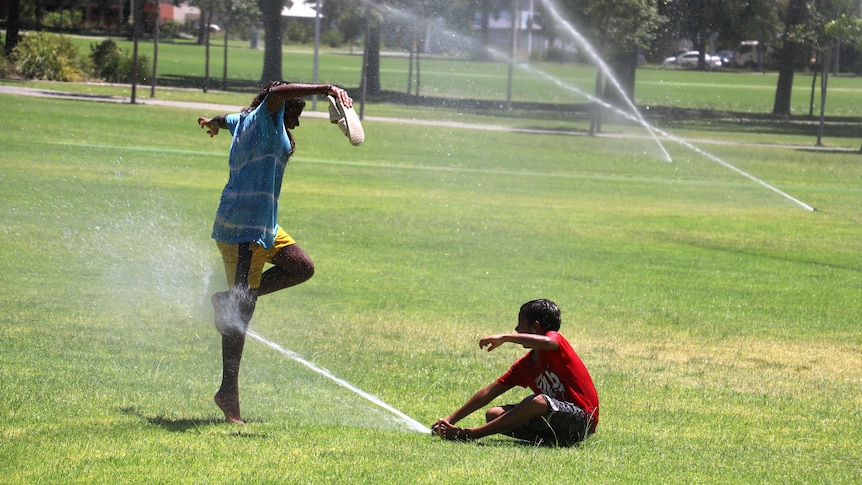 Two Indigenous children play under a sprinkler in a park, one seated and one standing with arms outstretched