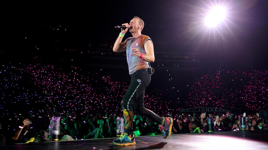 Singer Chris Martin on stage singing with a microphone in his hand and fans in the background