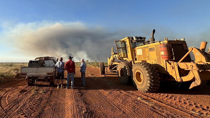 several station workers watching a large fire on the horizon, while standing next to a ute and a grader.