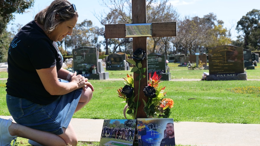 A woman kneels beside a cross that has photos and flowers on it.