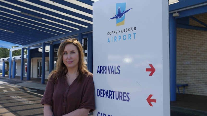 A Caucasian woman with dark brown hair and dress stands next to a board saying Coffs Harbour Airports departures, arrivals.