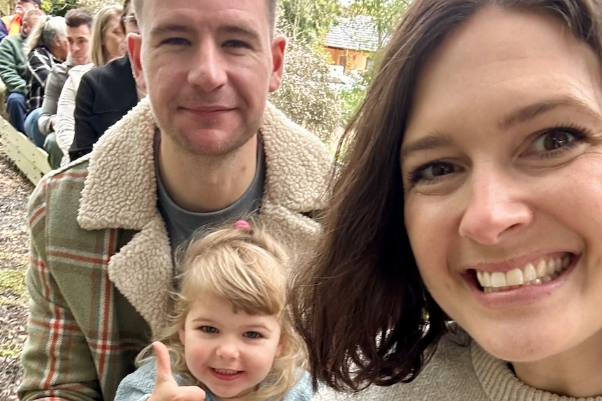 Rebecca Pritchard smiles while standing with her husband and daughter.