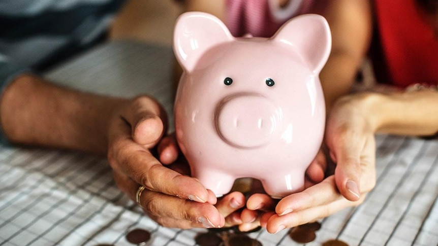 Two hands hold a light-pink ceramic piggy bank full of coins.