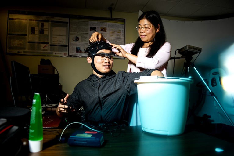 Yingzi Lin adjusts an EEG cap on the head of a volunteer as they plunge their arm into a white bucket