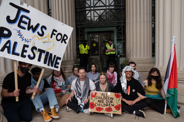 Jessica Metzger, wearing a face mask, sits among two rows of student protestors holding flags and signs.