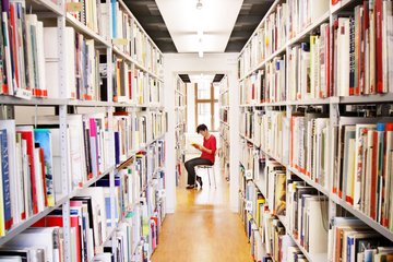 lesende Frau sitzt zwischen zwei deckenhohen Bücherregalen