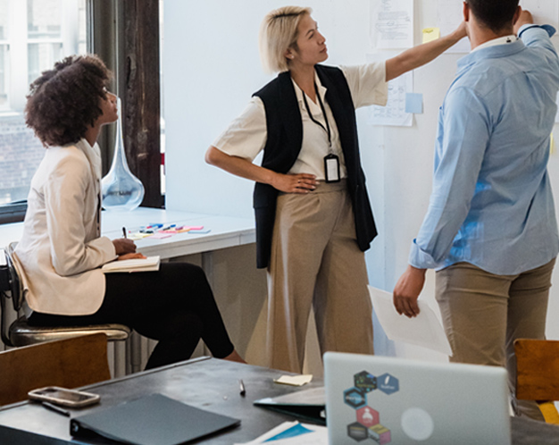 3 people in a meeting standing and point to paper on a wall and laptop with hex stickers on a nearby desk